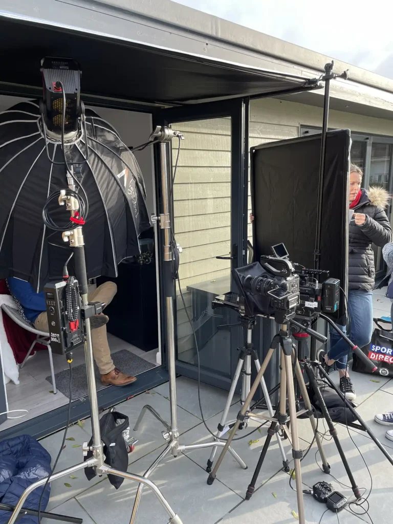 Behind the scenes of a video shoot showing high end cameras, lighting and microphones, along with crew members holding frames and reflectors.
