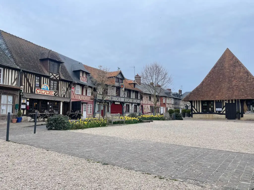 Timber-framed village square with creperie and steep-roofed hall under an overcast sky.
