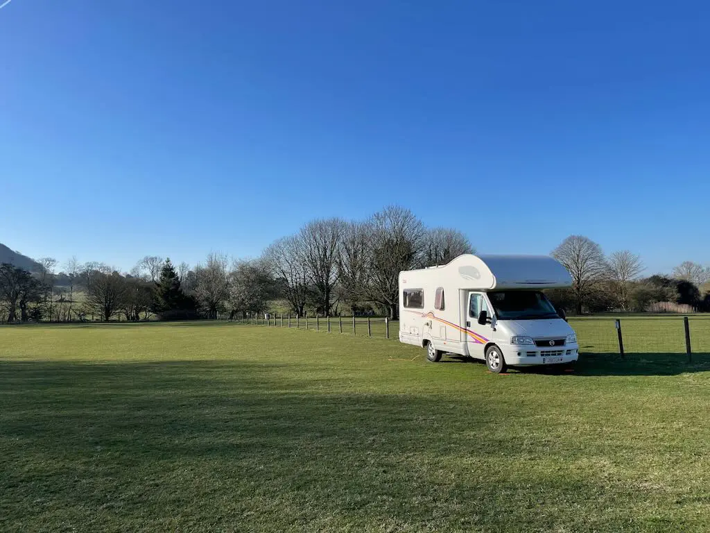 White motorhome parked on a wide grassy field under a bright blue sky, trees lining the background.
