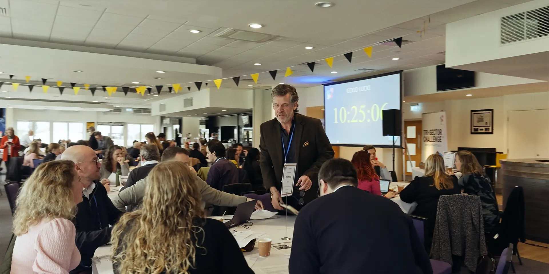 Still frame of an event video featuring business people at a conference, gathered around desks with a projector in the distance.